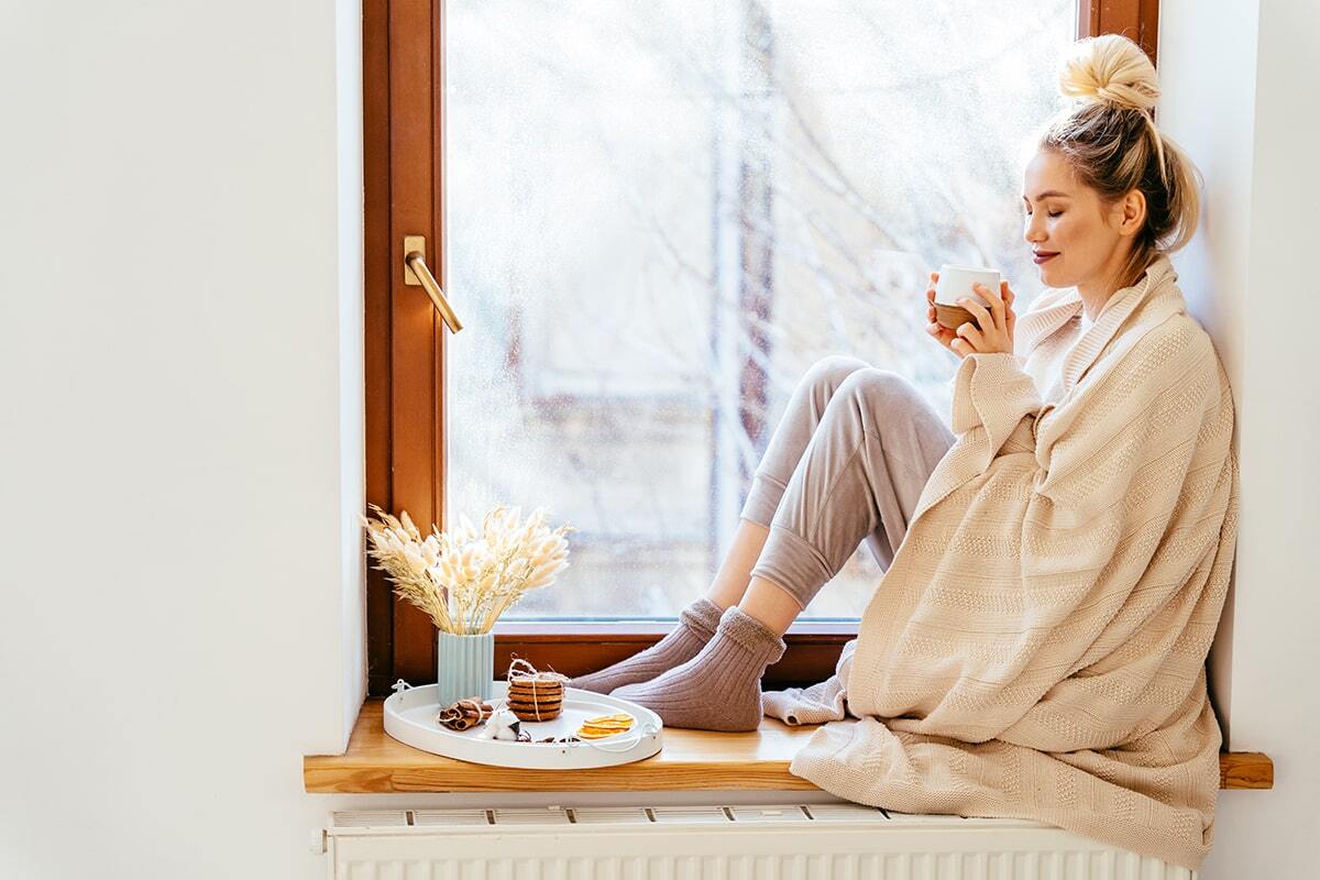 Giovane donna bionda seduta sul davanzale della finestra con in mano una tazza di tè avvolta in un plaid 